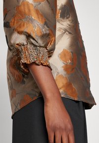 Détail de manche d'un blouse brillante à motif floral avec un poignet élastique sur le bras d'une personne, portée sur un pantalon noir.