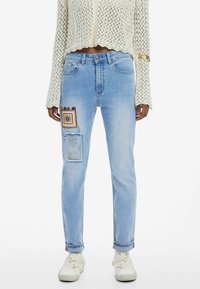 Jean en denim bleu clair avec une coupe décontractée, présentant des détails de patchwork artistique sur la cuisse gauche et un léger effet vieilli à l'ourlet.