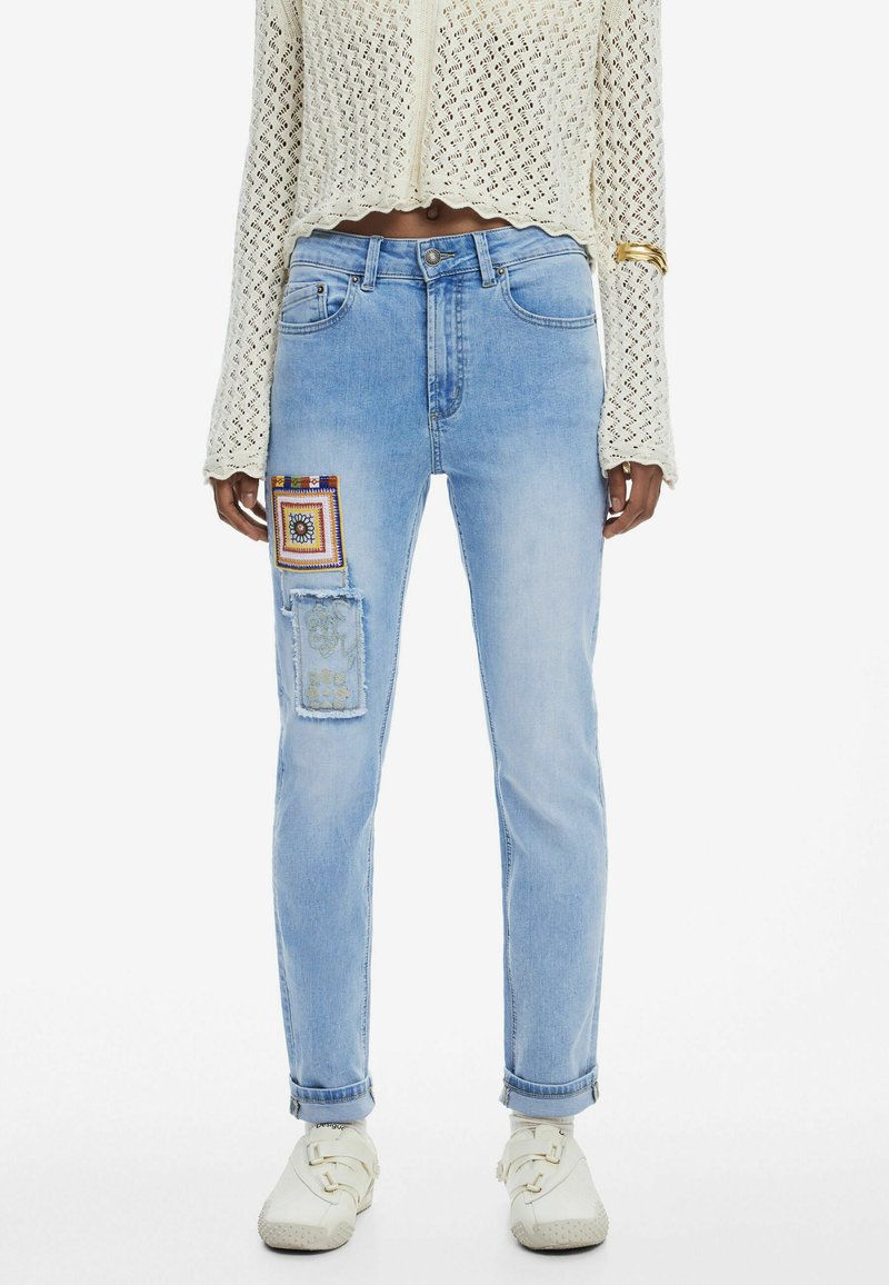 Jeans de mezclilla de color azul claro con un ajuste relajado, que presentan detalles artísticos de patchwork en el muslo izquierdo y un ligero desgastado en el dobladillo.