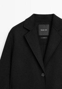 Czarny płaszcz z wełny z domieszkami, z wyciętym kołnierzem, zapinany na jeden guzik i o gładkiej fakturze. Metka widoczna wewnątrz w okolicy szyi.