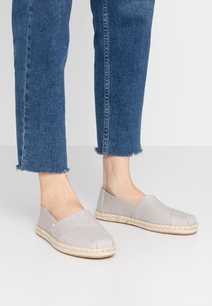 Fødder iført lysegrå slip-on espadrilles med flettede rebssåler, kombineret med blå, beskårne cowboybukser med flossede kanter.