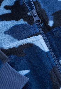 Gros plan d’un tissu polaire camouflage bleu avec une fermeture éclair bleu foncé et un poignet tricoté côtelé.