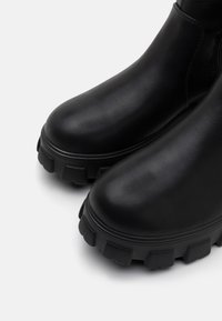 Fekete bőr bokacsizmák, texturált, vastag gumitalppal és lekerekített orral. A karcsú kialakítás minimalista varrásokkal és sima felülettel rendelkezik.