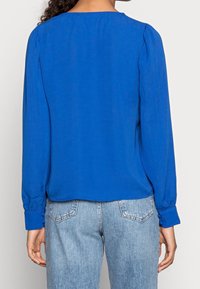 Blouse bleue à manches longues avec un col rond, texture douce et épaules froncées, associée à un jean délavé bleu clair et usé.