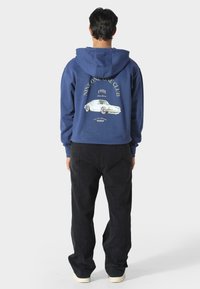 Felpa blu con cappuccio e lacci, con una grafica di un'auto e testo sul retro. Abbinata a pantaloni neri e scarpe di colore chiaro.