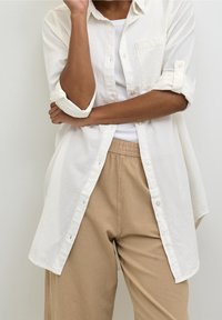 Chemise blanche à boutons en tissu léger, avec une poche et des manches retroussées, associée à un pantalon beige. Look simple et décontracté.