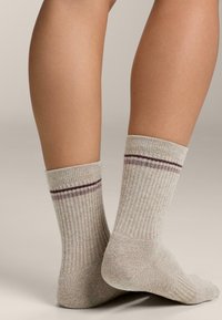 Nohy v béžových rebrovaných ponožkách s dvoma tenkými tmavofialovými pruhmi pri vrchu, jedna noha je na podlahe a druhá je mierne zdvihnutá na prstoch.