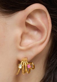Des boucles d'oreilles en or en forme de créole présentent trois anneaux entrelacés, accentués par une fleur en argent et une fleur rose, positionnées près d'une oreille.