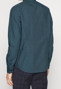 Langarmshirt van donkergroene stof met een herhalend lichtblauw geometrisch patroon. De overhemden hebben knoopmanchetten en een afgeronde zoom die het ontwerp afmaakt.