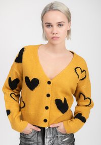 Jongere met kort blond haar, gekleed in een mosterdgele gebuttonde trui met zwarte hartpatronen en metallic broek.