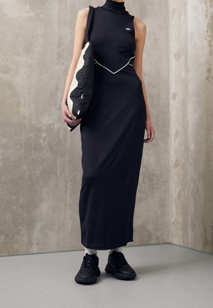 Schwarzes ärmelloses Kleid mit hohem Kragen, figurbetontem Design und kontrastierender weißer Paspelierung, kombiniert mit einer schwarz-weißen, gesteppte Tasche und schwarzen Sneakern.