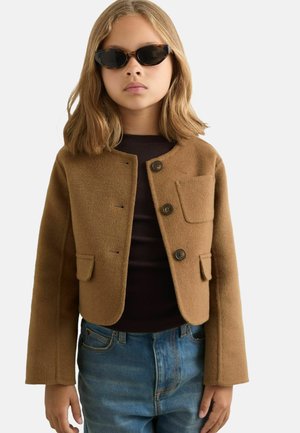 Braune Wolljacke mit runder Halslinie, Knopfverschluss und aufgesetzter Tasche. Über einem dunklen Oberteil getragen und mit blauen Jeans kombiniert.