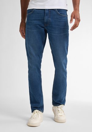 Jeans de mezclilla azul con un corte recto, diseño de cinco bolsillos y detalles de costura visibles; llevados con zapatillas de color claro.