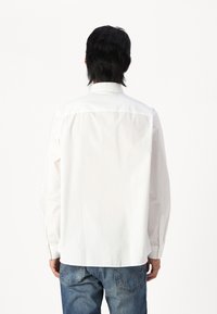 White long-sleeve shirt with a straight cut, buttoned cuffs, and a classic collar. Fabric appears smooth and lightweight. Worn with blue jeans.