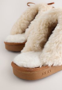 Paire de chaussons beige duveteux avec des semelles épaisses marron et une doublure en peluche douce, inclinée pour montrer la texture et la marque de la semelle.