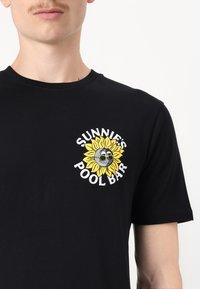 Svart bomullströja med en gul solrosgrafik med solglasögon och texten "SUNNIE'S POOL BAR" i vitt. Standard rund hals.