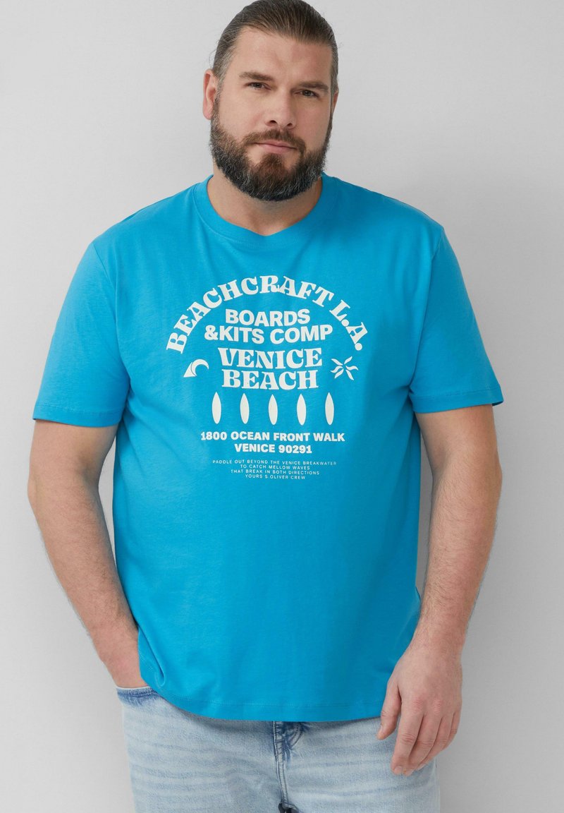 Turquoise t-shirt met korte mouwen met witte grafische tekst. Het shirt heeft een ronde halslijn en een relaxte fit, gecombineerd met lichte denim jeans.