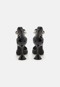 Sorte patent lær høyhælte sko med spiss tå, med glidelås bak og dekorative sølvdetaljer på hælen.