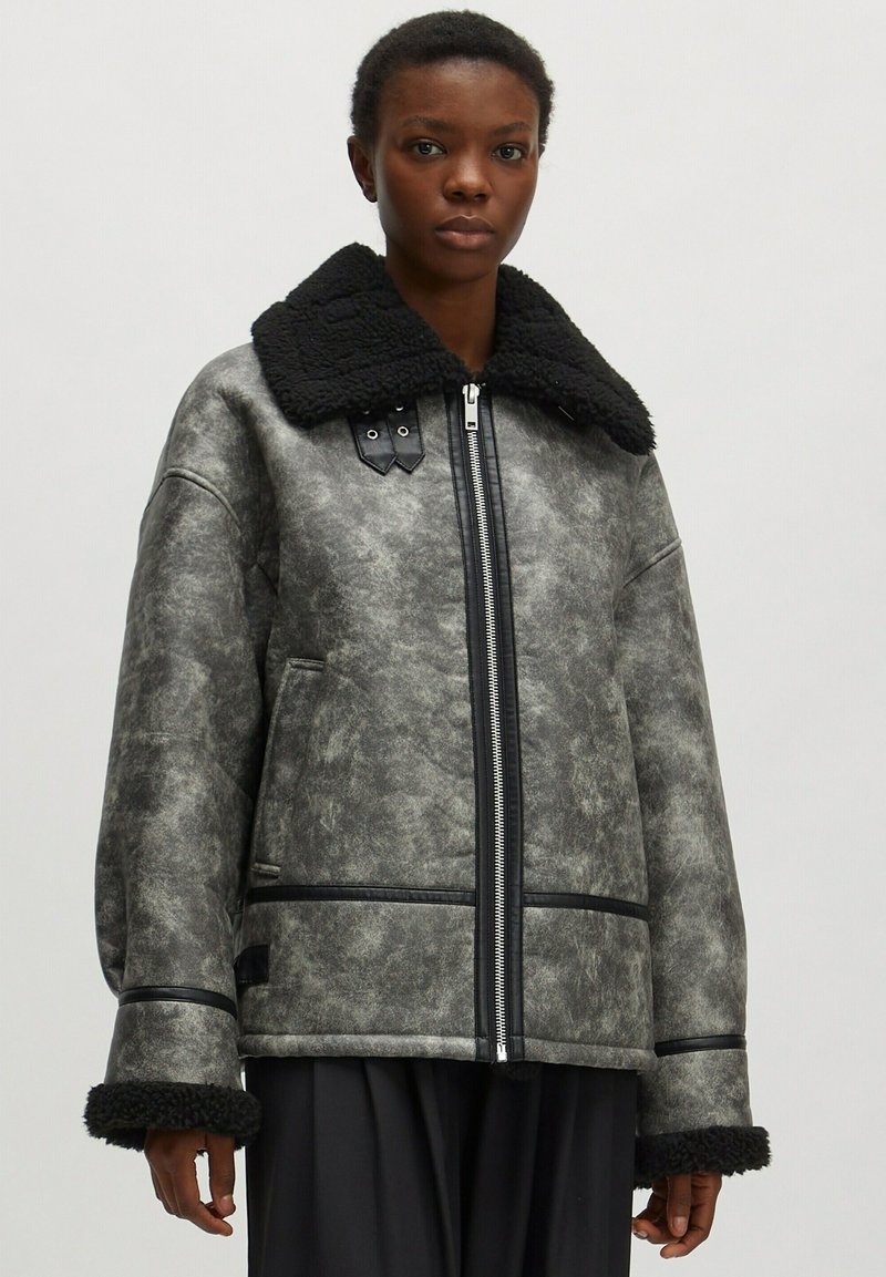 Graue Kunstlederjacke mit schwarzem Fellkragen, Reißverschluss und schwarzen Akzenten. Mit weit geschnittenen Ärmeln und einer strukturierten Oberfläche.