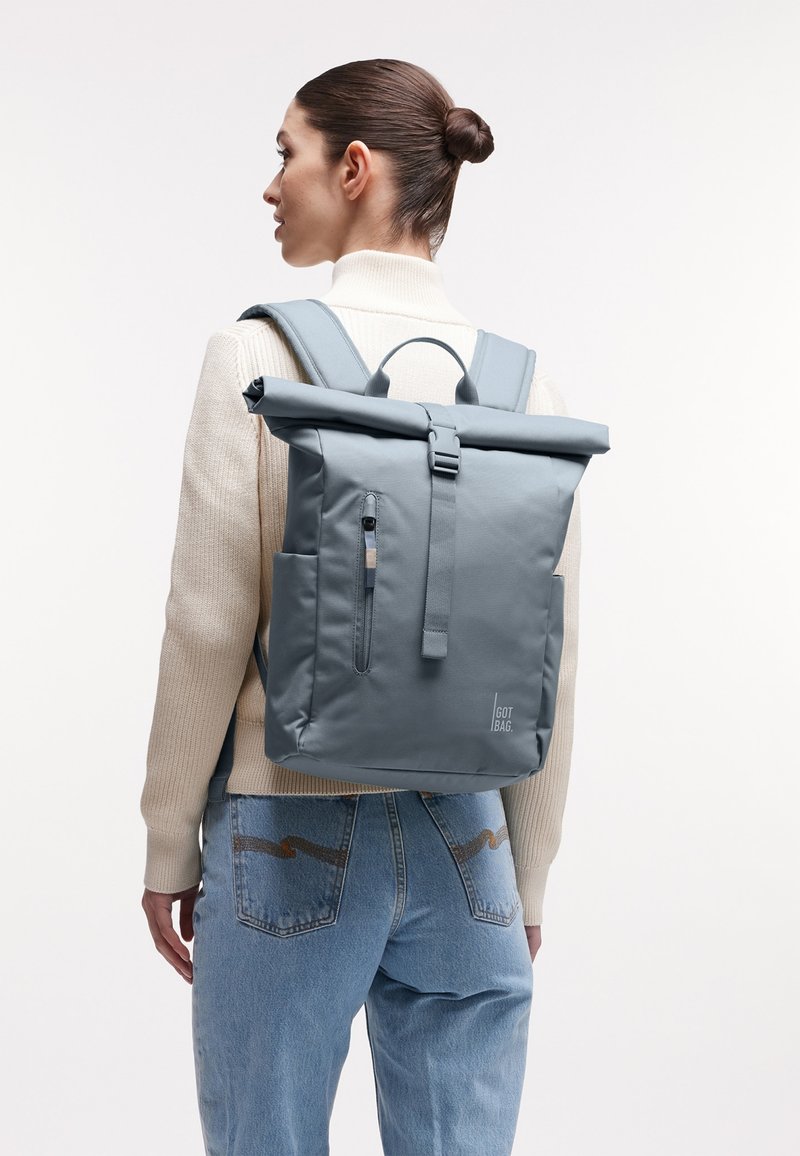 Mujer con cabello castaño recogido en un moño, vestida con un suéter beige y jeans azules, lleva una mochila gris con tapa enrollable y bolsillo frontal con cremallera.