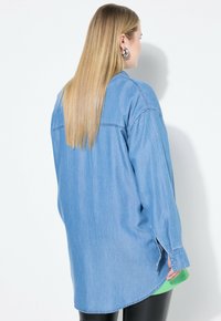 Camicia di jeans a maniche lunghe di colore azzurro, vestibilità rilassata, con spacchi laterali, polsini con bottone e orlo arrotondato. Indossata sopra un top verde.