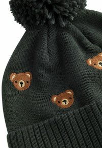 Strikket mørkegrøn hue med ribbet manchet. Har broderede bjørneansigtter i brunt, med en fluffy pompon på toppen. Blød tekstur.