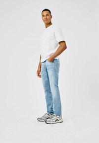 Hombre de pie con la mano derecha en el bolsillo, vistiendo una camiseta blanca, jeans de mezclilla azul claro y zapatillas grises sobre un fondo blanco liso.