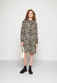 Robe imprimée à manches longues avec décolleté en trou de serrure, présentant un motif vert et noir. Complétée par un sac à main blanc et des chaussures noires épaisses.