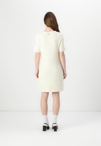 Robe en crème tissée avec des manches courtes, motif texturé, détail dans le dos, et une silhouette droite, associée à des chaussures à talons carrés noires.