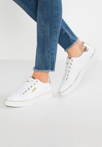 Baskets blanches avec une surface texturée, accents dorés sur le talon et les côtés, et lacets blancs. Associées à un jean bleu cropped.