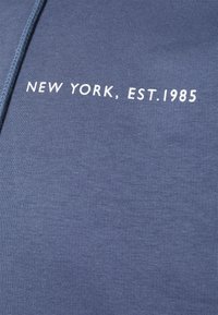 Blått tyg med vit text tryckt "NEW YORK, EST. 1985" och en flätad blå dragsnöre synlig på vänster sida.