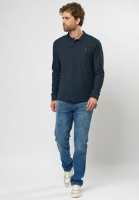 Marineblå langærmet polo med krave og en lille rød logo. Kombineret med lyseblå jeans og hvide sneakers.