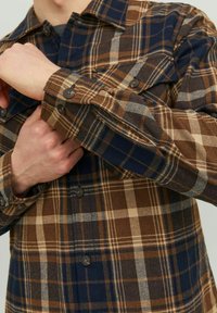 Camisa a cuadros marrón y azul marino con puños abotonados, que presenta una textura en la tela y un gran patrón de cuadros. Incluye bolsillos en el pecho y detalles en el cuello.