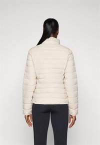 Pufferjack in lichtbeige met een quiltpatroon, hoge kraag en wijde mouwen. Gladde textuur, gedragen over zwarte leggings, weergegeven van achteren.