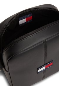 Μαύρο δερμάτινο τσαντάκι χιαστί με φερμουάρ, διακοσμημένο με λογότυπο σε κόκκινο, λευκό και μπλε στη μπροστινή πλευρά. Το εσωτερικό είναι επενδυμένο.