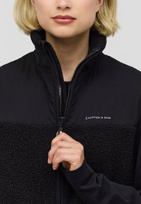 Schwarwollweste aus Fleece und strukturiertem Stoff mit Reißverschluss. Mit hohem Kragen und Logo auf der Brust. Hand in der Nähe des Reißverschlusses positioniert.