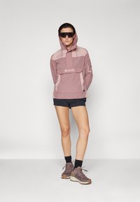 Sudadera con capucha de color rosa claro, con un bolsillo frontal y logo, combinada con pantalones cortos negros. El modelo usa gafas de sol y zapatos de senderismo marrones. Textura suave.