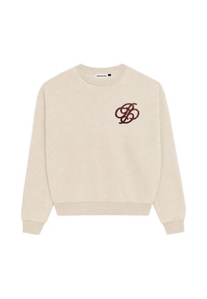 Mann mit kurzen Haaren und Bart, der einen beigen Pullover mit dunkelrotem Monogramm auf der Brust und schwarze Hosen trägt, schaut nach links.