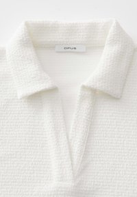 Polo bianco caratterizzato da un tessuto strutturato con un motivo intrecciato, un colletto morbido e ripiegabile, e un'etichetta con la scritta "OPUS" sul collo.
