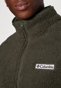 Olive zielona bluza polarowa z wysokim kołnierzem, zamek błyskawiczny oraz biała naszywka z logo Columbia na piersi. Miękki, teksturowany materiał.