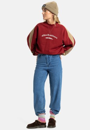 Červená a béžová oversized mikina s logom, vysoké modré džínsy, mnohofarebné pletené ponožky a tmavohnedé slip-on topánky.