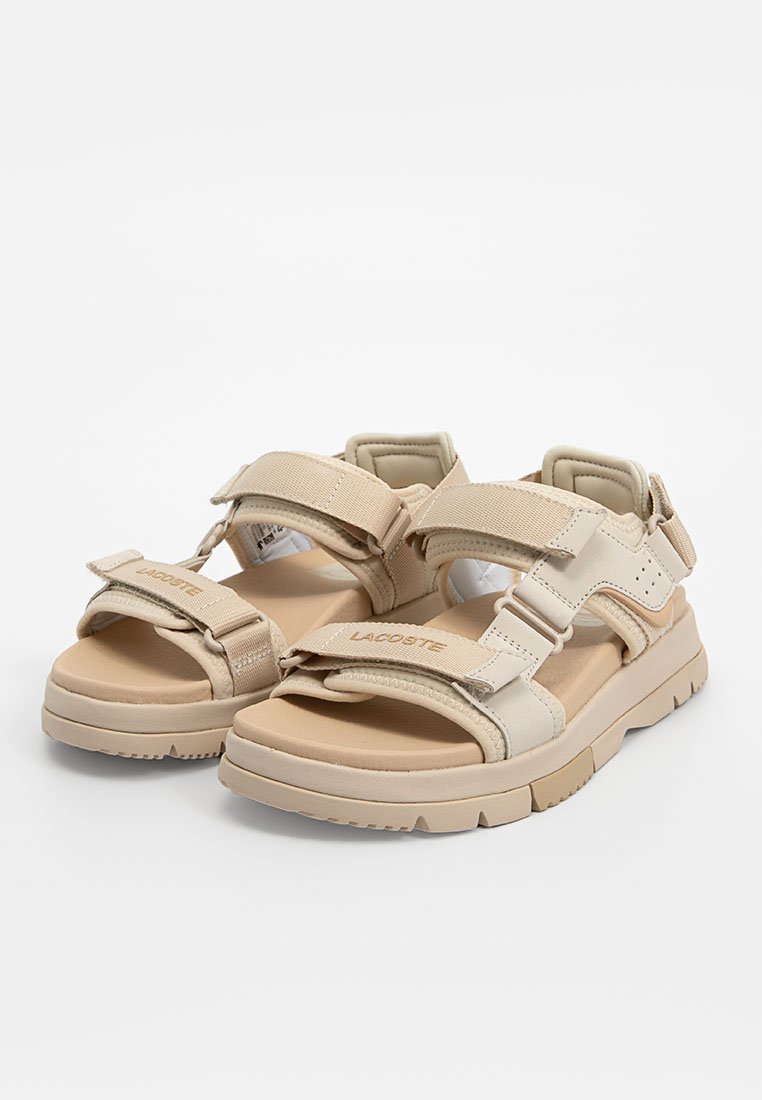 Sandales beiges avec sangles réglables en Velcro, semelle en caoutchouc épaisse, semelle rembourrée et logo en relief sur la sangle. Design structuré pour le confort.
