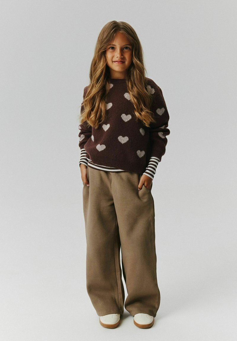 Jeune fille souriante portant un pull marron à motif cœur, des manches de chemise rayées, un pantalon large beige et des baskets blanches, les mains dans les poches.