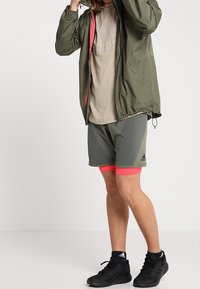 Chaqueta ligera verde oliva con forro rosa, camiseta color beige, pantalones cortos grises con acentos rosas y zapatillas deportivas negras.
