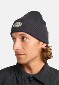 Gorro de punto negro con canalé y una parte superior redondeada, adornado con un parche de logo en crema en la parte delantera. Usado por una persona con cabello claro y una chaqueta oscura.