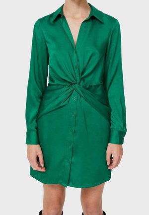 Vrouw die een groen, lang mouwen, knoopsluitingsjurk draagt met een gedraaide knoopdetail ter hoogte van de taille, staand tegen een effen achtergrond.