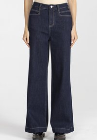 Kobieta nosząca ciemnoniebieskie dżinsy z wysokim stanem i szerokimi nogawkami, z kontrastowymi szwami oraz czarnymi butami na platformie, stojąca na jednolitym tle.