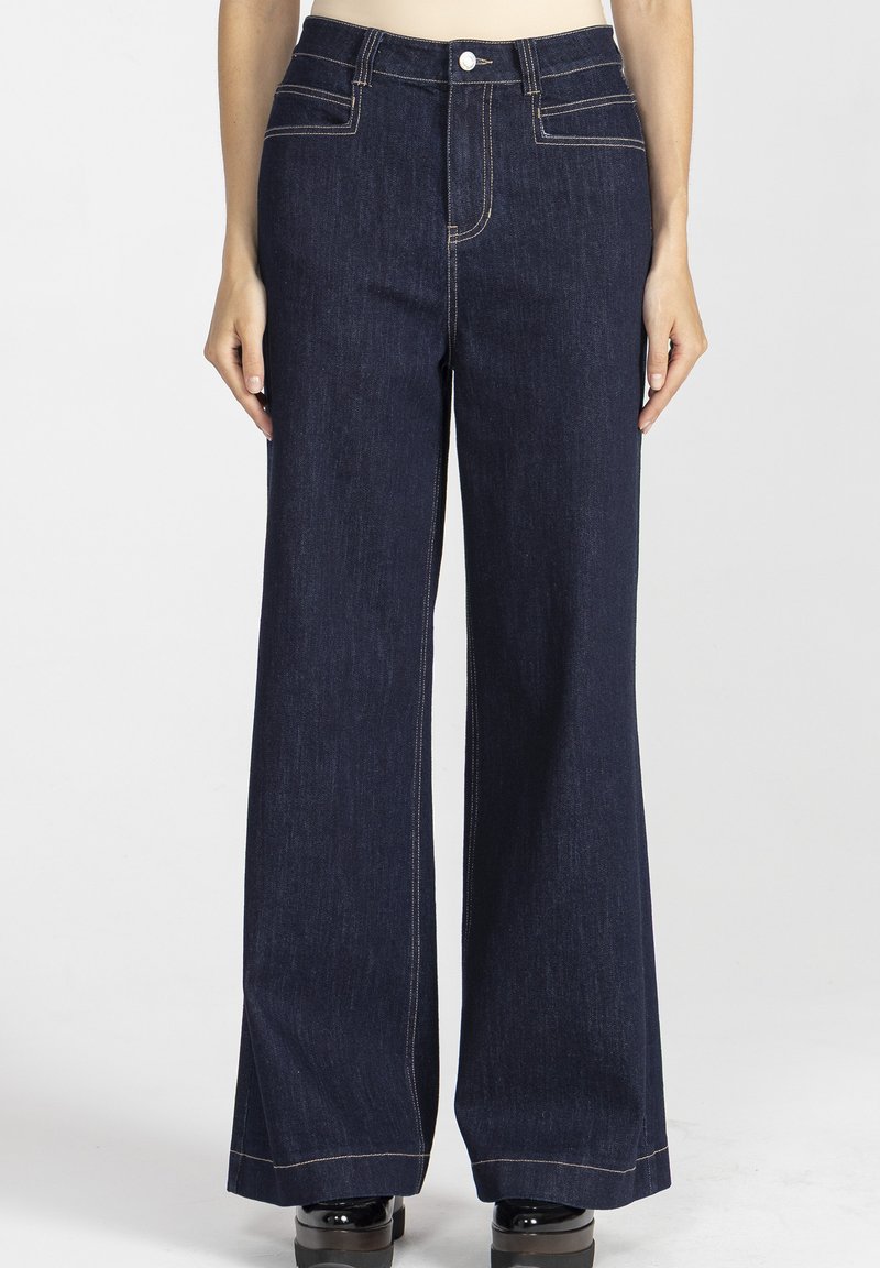 Kobieta nosząca ciemnoniebieskie dżinsy z wysokim stanem i szerokimi nogawkami, z kontrastowymi szwami oraz czarnymi butami na platformie, stojąca na jednolitym tle.