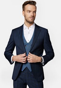 Granatowy, taliowany garnitur z wytłaczanym kamizelką w kratkę i białą koszulą, z guzikami dekoracyjnymi oraz wyraźnie zaznaczonymi ramionami.