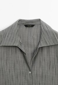 Camicia grigia con colletto caratterizzata da strisce verticali di diverse larghezze, con una texture liscia e una chiusura a bottone singola al collo.
